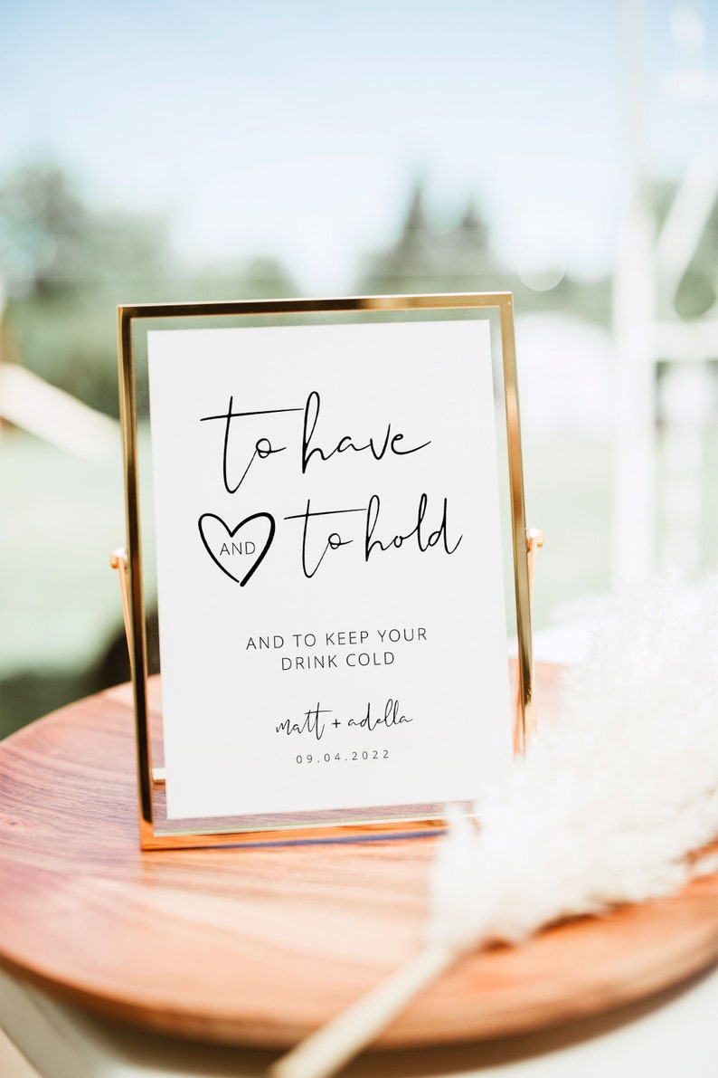 May include: A gold-framed sign with the text "to have and to hold" and a heart symbol. Below the text is "AND TO KEEP YOUR DRINK COLD" and the names "matt + adella" and the date "09.04.2022".
