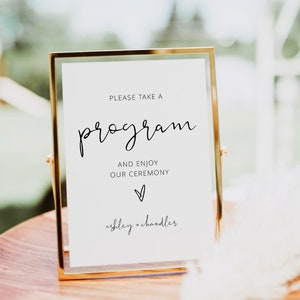 May include: A gold-framed sign with the words "PLEASE TAKE A PROGRAM" in elegant script. Below, it reads "AND ENJOY OUR CEREMONY" with a heart symbol and the names "ashley + chandler". The sign is on a wooden surface.