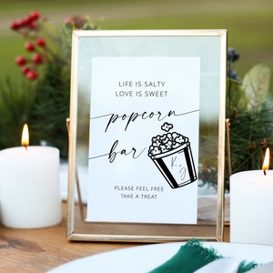 May include: A gold-framed sign with a white background and black text that reads "Life is salty, love is sweet, popcorn bar". A black and white illustration of a popcorn bucket with the initials "K.J." is below the text. The sign is on a wooden table with two white candles and a green napkin.