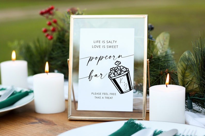 May include: A gold-framed sign with a white background and black text that reads "Life is salty, love is sweet, popcorn bar". A black and white illustration of a popcorn bucket with the initials "K.J." is below the text. The sign is on a wooden table with two white candles and a green napkin.