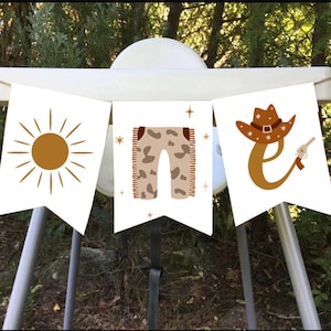 May include: A banner with three white pennants featuring a sun, pants, and a cowboy hat with a gun. The sun is brown with radiating lines. The pants are tan with brown spots. The cowboy hat is brown with a silver buckle.