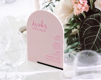 May include: A pink and white arch-shaped sign with the text "drinks with Chloe" in pink lettering. The sign lists various beer, spirits, and wine options, including Amstel Light, Budweiser, Absolut Vodka, Gordon's Gin, Bacardi Rum, Cabernet Sauvignon, and Chardonnay. The sign also features a signature drink called "The French Kiss" with whiskey, lemon juice, and lemon soda.