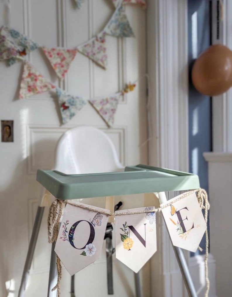 Puede incluir: Una silla alta blanca con una bandeja verde y una pancarta que dice "ONE" en un dise&ntilde;o floral. La pancarta est&aacute; hecha de tela beige con ilustraciones florales y de mariposas. La pancarta est&aacute; atada con un cordel.
