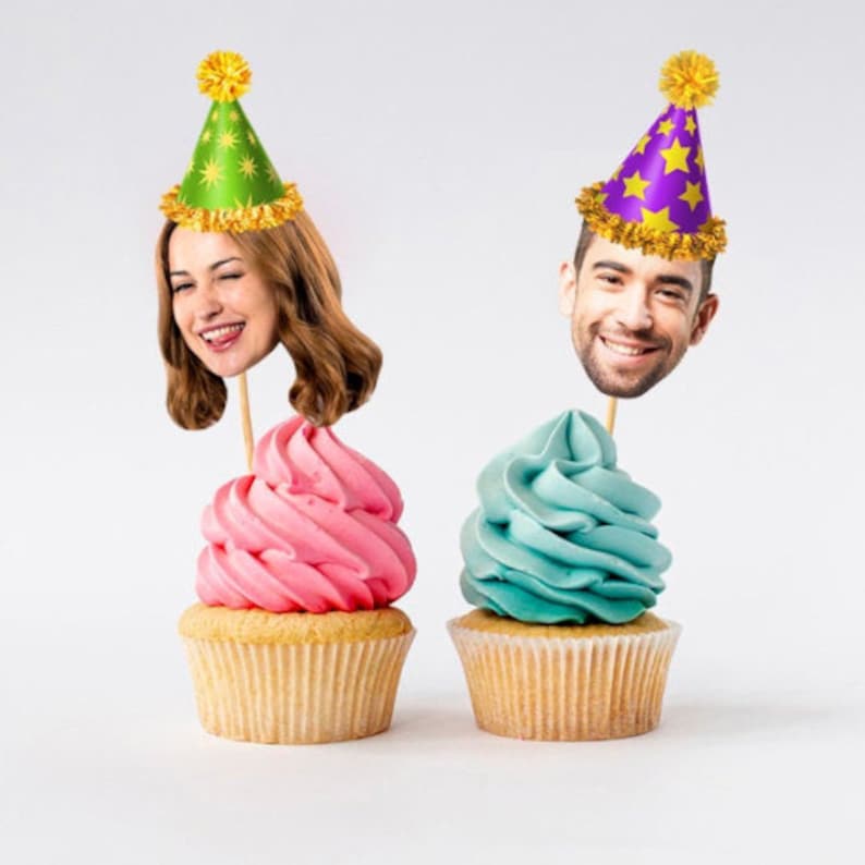 May include: Two cupcakes with frosting, one pink and one blue, topped with a person's face on a stick wearing a party hat. The hats are green and purple with gold stars.
