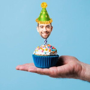 May include: A blue cupcake with white icing and colourful sprinkles topped with a green and yellow party hat and a lit candle. The hat has a man's face with a smile.