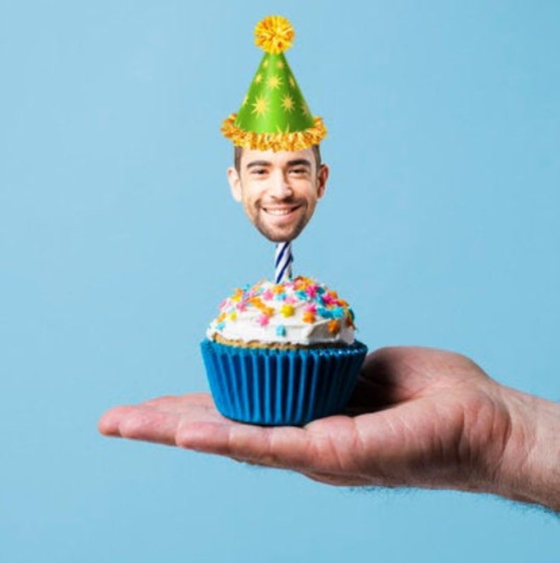 May include: A blue cupcake with white icing and colourful sprinkles topped with a green and yellow party hat and a lit candle. The hat has a man's face with a smile.