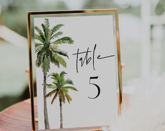 May include: Gold framed table number card with a white background and two watercolor palm trees. The text "table 5" is written in black cursive.
