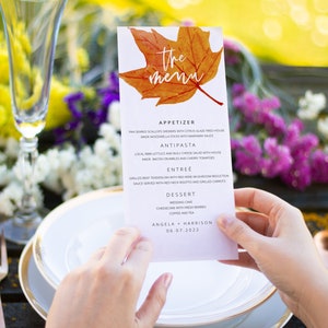 May include: A wedding menu with a fall theme. The menu features a watercolor illustration of a maple leaf in shades of orange and yellow. The menu lists the appetizers, antipasto, entree, and dessert. The text at the bottom of the menu reads "Angela + Harrison 06.07.2022".