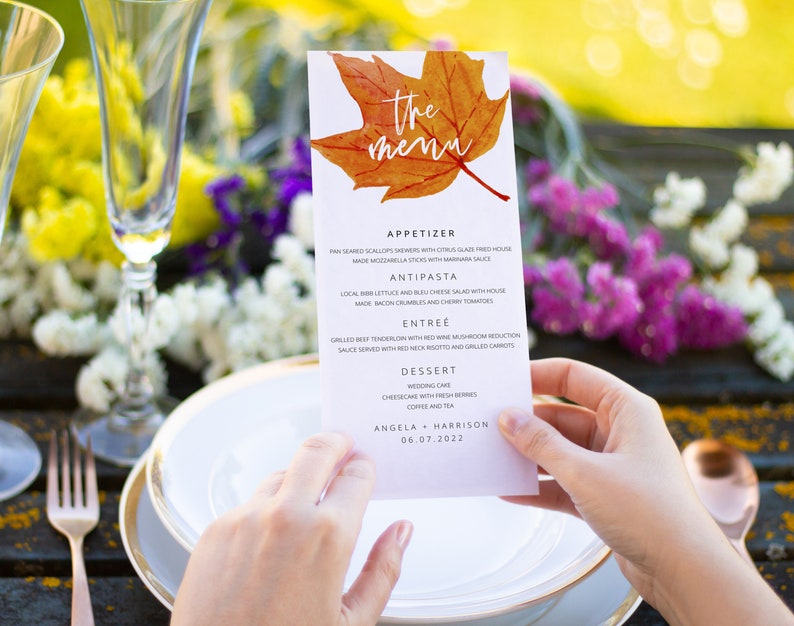May include: A wedding menu with a fall theme. The menu features a watercolor illustration of a maple leaf in shades of orange and yellow. The menu lists the appetizers, antipasto, entree, and dessert. The text at the bottom of the menu reads "Angela + Harrison 06.07.2022".