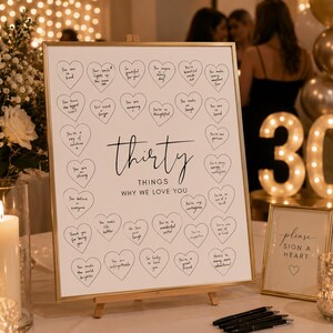 May include: A white rectangular sign in a gold frame featuring the text 'thirty THINGS WHY WE LOVE YOU' surrounded by thirty hand-drawn hearts containing handwritten messages. A smaller sign next to it reads 'please SIGN A HEART'.