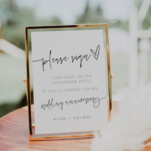 May include: A gold-framed sign with a white background and black text that reads "Please sign your name on the champagne bottle to help us celebrate our first wedding anniversary, Aline + Kaique".
