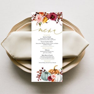 May include: A rectangular menu card with floral and pumpkin illustrations, placed on a plate with a folded napkin. The menu features a gold script font that reads "menu". The card's design includes autumn colors.