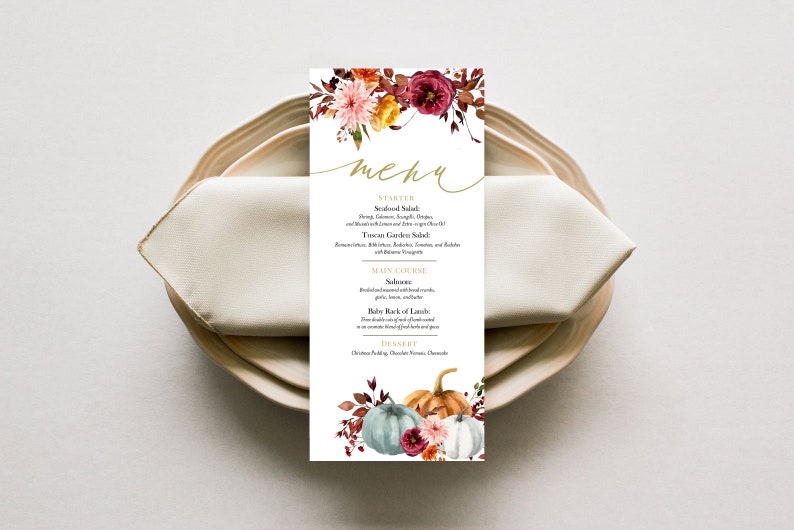 May include: A rectangular menu card with floral and pumpkin illustrations, placed on a plate with a folded napkin. The menu features a gold script font that reads "menu". The card's design includes autumn colors.