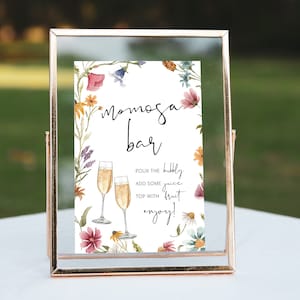May include: A printable sign with a floral border and the text "Mimosa Bar". The sign instructs guests to "Pour the bubbly, add some juice, top with fruit, enjoy!"