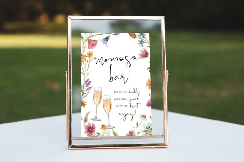 May include: A printable sign with a floral border and the text "Mimosa Bar". The sign instructs guests to "Pour the bubbly, add some juice, top with fruit, enjoy!"
