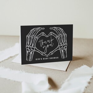 May include: Black thank you card with white skeleton hands forming a heart shape. The text inside the heart reads "thank you". The bottom of the card reads "Kim's Baby Shower".