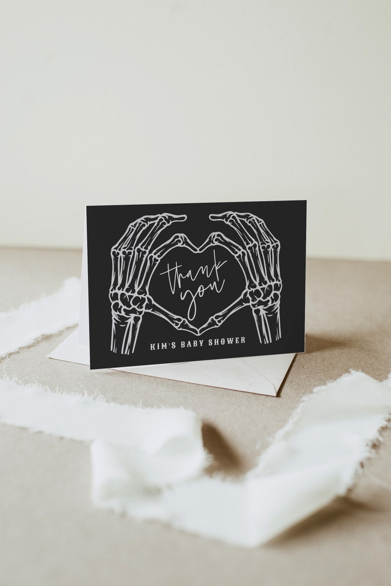 May include: Black thank you card with white skeleton hands forming a heart shape. The text inside the heart reads "thank you". The bottom of the card reads "Kim's Baby Shower".