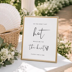 以下が含まれることがあります： 金色のフレームに入った看板には、黒い文字で「SO YOU DON'T GET hot WHILE WE TIE the knot PLEASE TAKE A FAN」と書かれており、白い折りたたみ扇子が入った編みかごと、白いバラと緑の葉のブーケの隣に置かれています。