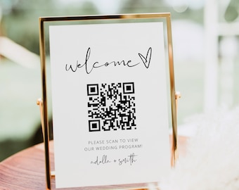 Op de afbeelding: Een goudkleurig omlijst bord met het woord "welcome" in cursief en een hartsymbool. Hieronder staat een QR-code en de tekst "PLEASE SCAN TO VIEW OUR WEDDING PROGRAM!" met de namen "adella + smith". Het bord staat op een houten oppervlak.