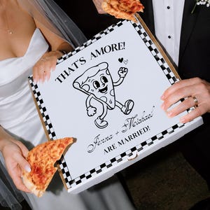 Könnte beinhalten: Ein weißer, quadratischer Pizzakarton mit schwarz-weißem Schachbrettmuster am Rand. Der Deckel zeigt eine Comic-Pizzastück-Figur und den Text THAT'S AMORE! Jenna + Michael ARE MARRIED! Der Karton wird von zwei Personen in festlicher Kleidung gehalten.