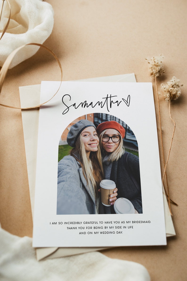 May include: A white card with a photo of two women smiling at the camera. The card has the text "Samantha" with a heart symbol and the message "I am so incredibly grateful to have you as my bridesmaid. Thank you for being by my side in life and on my wedding day."