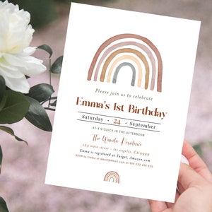 May include: A white invitation card with a watercolor rainbow design in shades of brown, beige, and green. The text reads "Emma's 1st Birthday" with event details. The card is held in a person's hand.