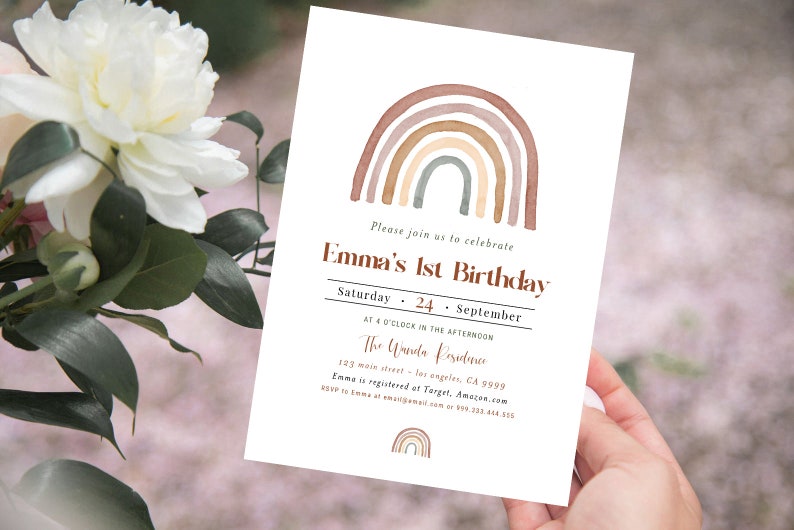 May include: A white invitation card with a watercolor rainbow design in shades of brown, beige, and green. The text reads "Emma's 1st Birthday" with event details. The card is held in a person's hand.