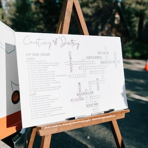May include: A white crossword puzzle on an easel with the title "Courtney & Dmitry Sip and Solve". The crossword puzzle has clues about the couple's relationship and wedding.