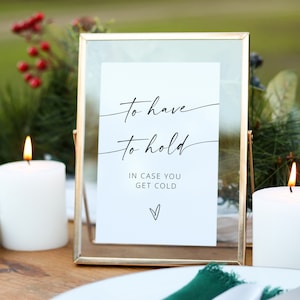 May include: A gold-framed sign with the text "To have to hold in case you get cold" and a small heart at the bottom. The sign is sitting on a wooden table with two white candles and green napkins.