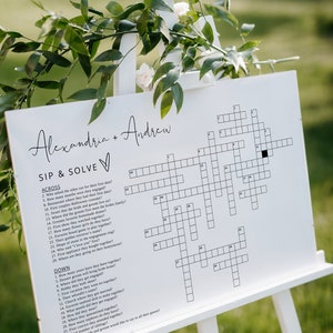 May include: A white sign with a crossword puzzle on it. The sign is titled "Sip & Solve" and has the names Alexandria and Andrew at the top. The crossword puzzle has clues about the couple's relationship.