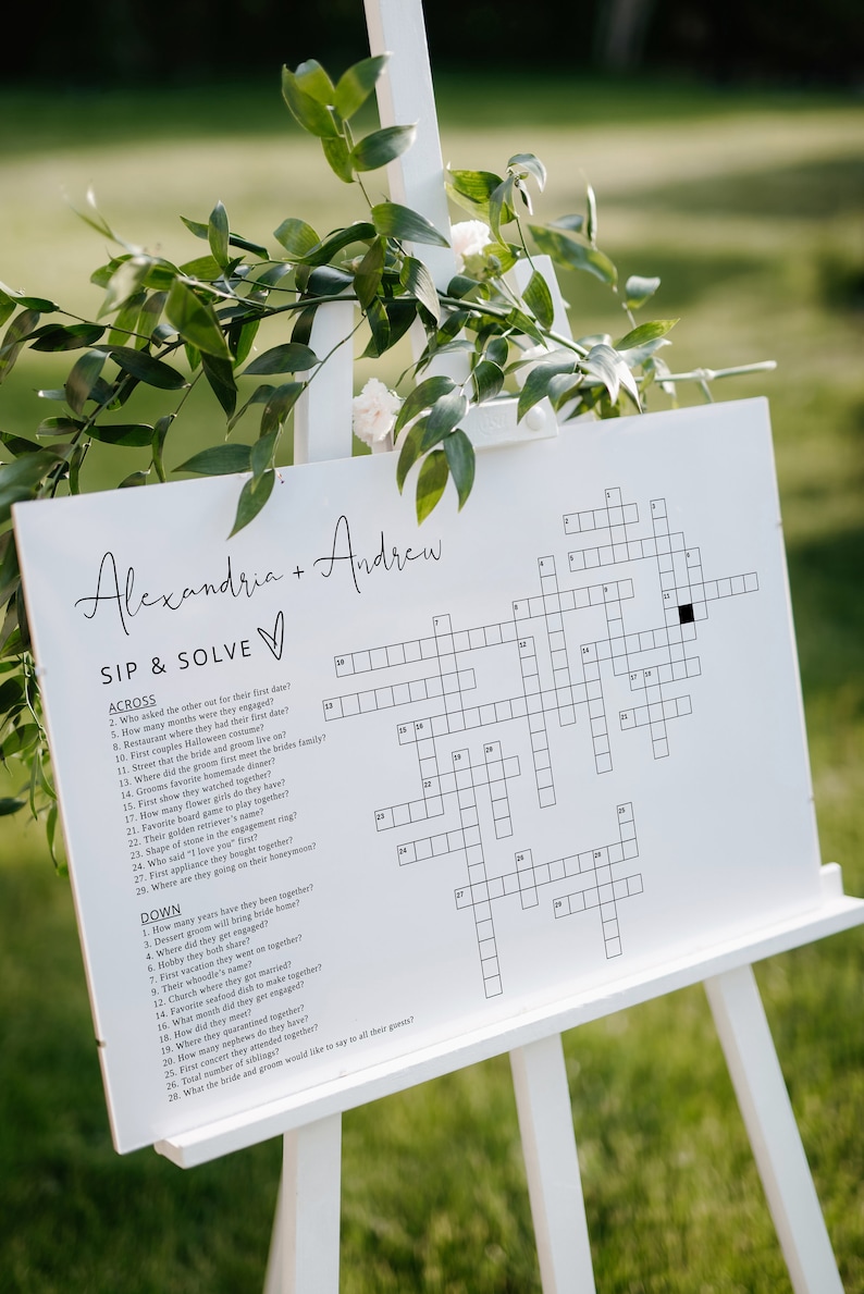 May include: A white sign with a crossword puzzle on it. The sign is titled "Sip & Solve" and has the names Alexandria and Andrew at the top. The crossword puzzle has clues about the couple's relationship.