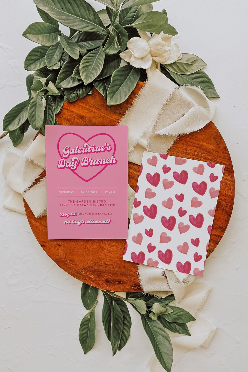 May include: Pink invitation card with the text "Galentine's Day Brunch" in a retro font. The card also includes the date, time, location, and RSVP information. The card is on a wooden surface with a white ribbon and green leaves.