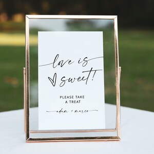 May include: A gold-framed glass sign with the text "love is sweet!" and "Please take a treat - adam + marco" on a white background.