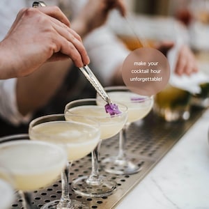 May include: A bartender garnishes cocktails with purple flowers using tongs. The image shows a row of coupe glasses filled with a light-colored drink. Text in the image reads: "make your cocktail hour unforgettable!"