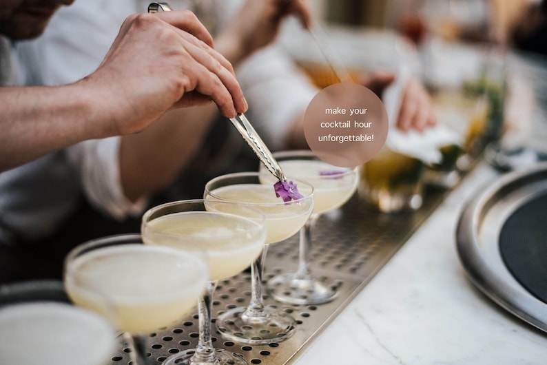May include: A bartender garnishes cocktails with purple flowers using tongs. The image shows a row of coupe glasses filled with a light-colored drink. Text in the image reads: "make your cocktail hour unforgettable!"