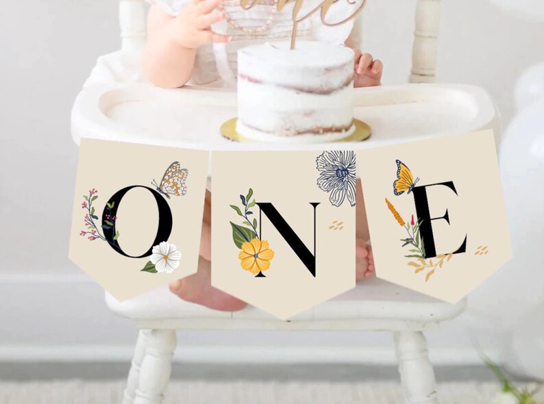 Puede incluir: Una silla alta beige con una pancarta floral que dice "ONE" en letras negras. La pancarta tiene mariposas, flores y hojas en tonos amarillos, azules y verdes. Un beb&eacute; est&aacute; sentado en la silla alta con un pastel delante de &eacute;l.