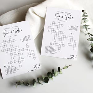 May include: Two white crossword puzzle cards with the text "Jessica + Michael Sip & Solve 10.04.2024" in black script. The cards have a crossword grid and clues. A sprig of greenery and a white cloth are in the background.