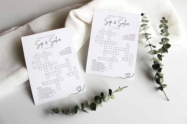 May include: Two white crossword puzzle cards with the text "Jessica + Michael Sip & Solve 10.04.2024" in black script. The cards have a crossword grid and clues. A sprig of greenery and a white cloth are in the background.