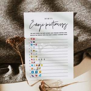 May include: A white card with the text "DIRTY Emoji Pictionary" printed in black. The card has a list of numbers 1 through 13 with emojis next to each number. The card is on a brown surface with a dried flower.