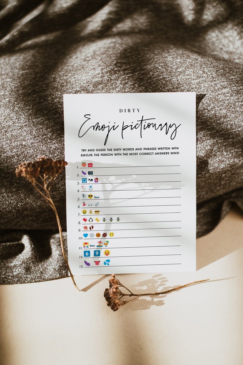 May include: A white card with the text "DIRTY Emoji Pictionary" printed in black. The card has a list of numbers 1 through 13 with emojis next to each number. The card is on a brown surface with a dried flower.