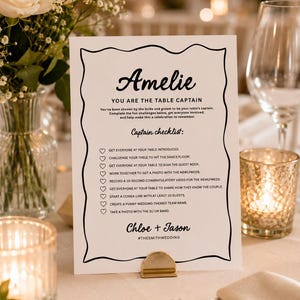 Op de afbeelding: Een witte rechthoekige tafelkaart voor een bruiloft met een zwarte decoratieve rand. De tekst bevat Amelie, You Are The Table Captain, Captain checklist, een takenlijst met hartjes, en eindigt met Chloe + Jason en #THESMITHWEDDING.