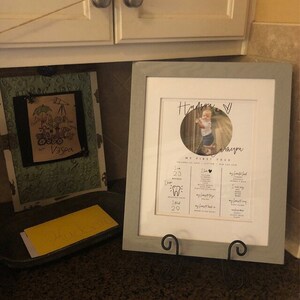 May include: A framed photo of a baby girl with a gray frame. The photo is a close-up of the baby girl and includes text that says "My First Year" and lists milestones such as age, weight, and teeth. The baby's name is "Hudson" and the photo is on a gray stand.