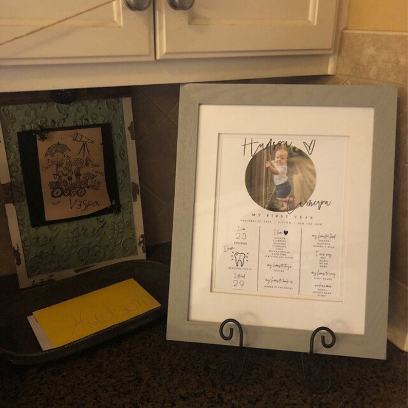 May include: A framed photo of a baby girl with a gray frame. The photo is a close-up of the baby girl and includes text that says "My First Year" and lists milestones such as age, weight, and teeth. The baby's name is "Hudson" and the photo is on a gray stand.