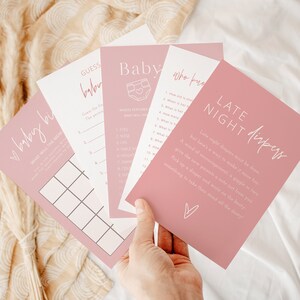 以下が含まれることがあります： ピンクのベビーシャワーゲームカードのセットで、白いテキストが書かれています。カードには、「ベビービンゴ」、「ベビーを一番よく知っているのは誰」、「ベビーフードを当てよう」、「深夜のおむつ」などのゲームが含まれています。