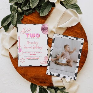May include: A pink and white birthday invitation with a cow print border. The invitation says "Two" in pink letters and "Amanda's Second Birthday!" in pink and white letters. The invitation also includes a guitar, a cowboy hat, a star, and a cowboy boot. The invitation is on a wooden surface with a white ribbon and green leaves.