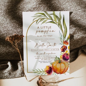 Pu&ograve; includere: Invito per baby shower con zucca e design floreale ad acquerello. Il testo recita "A Little Pumpkin is on the way!" con i dettagli dell'evento. Include una cornice dorata e elementi decorativi.