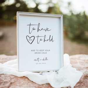 May include: White sign with black text that reads "to have and to hold and to keep your drink cold matt + adella 09.04.2022". The sign is sitting on a rock with a white cloth draped over it.