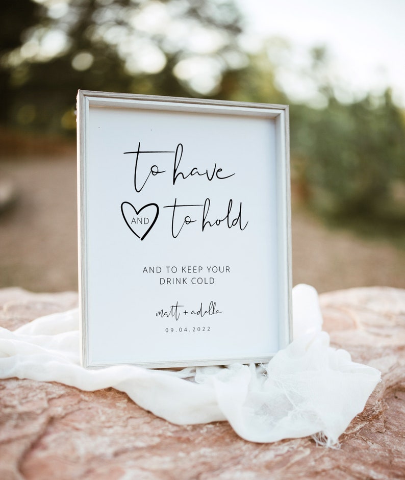 May include: White sign with black text that reads "to have and to hold and to keep your drink cold matt + adella 09.04.2022". The sign is sitting on a rock with a white cloth draped over it.