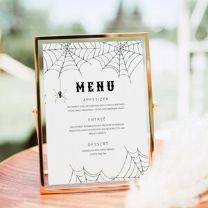 May include: A gold-framed menu with a white background and black spiderweb illustrations. The menu lists "MENU", "APPETIZER", "PAN SEARED SCALLOPS SKEWERS WITH CITRUS GLAZE FRIED", "HOUSE MADE MOZZARELLA STICKS WITH MARINARA SAUCE", "ENTRE&Eacute;", "GRILLED BEEF TENDERLOIN WITH RED WINE MUSHROOM", "REDUCTION SAUCE SERVED WITH RED NECK RISOTTO AND", "GRILLED CARROTS", "DESSERT", "CHEESECAKE WITH FRESH BERRIES", "COFFEE AND TEA".