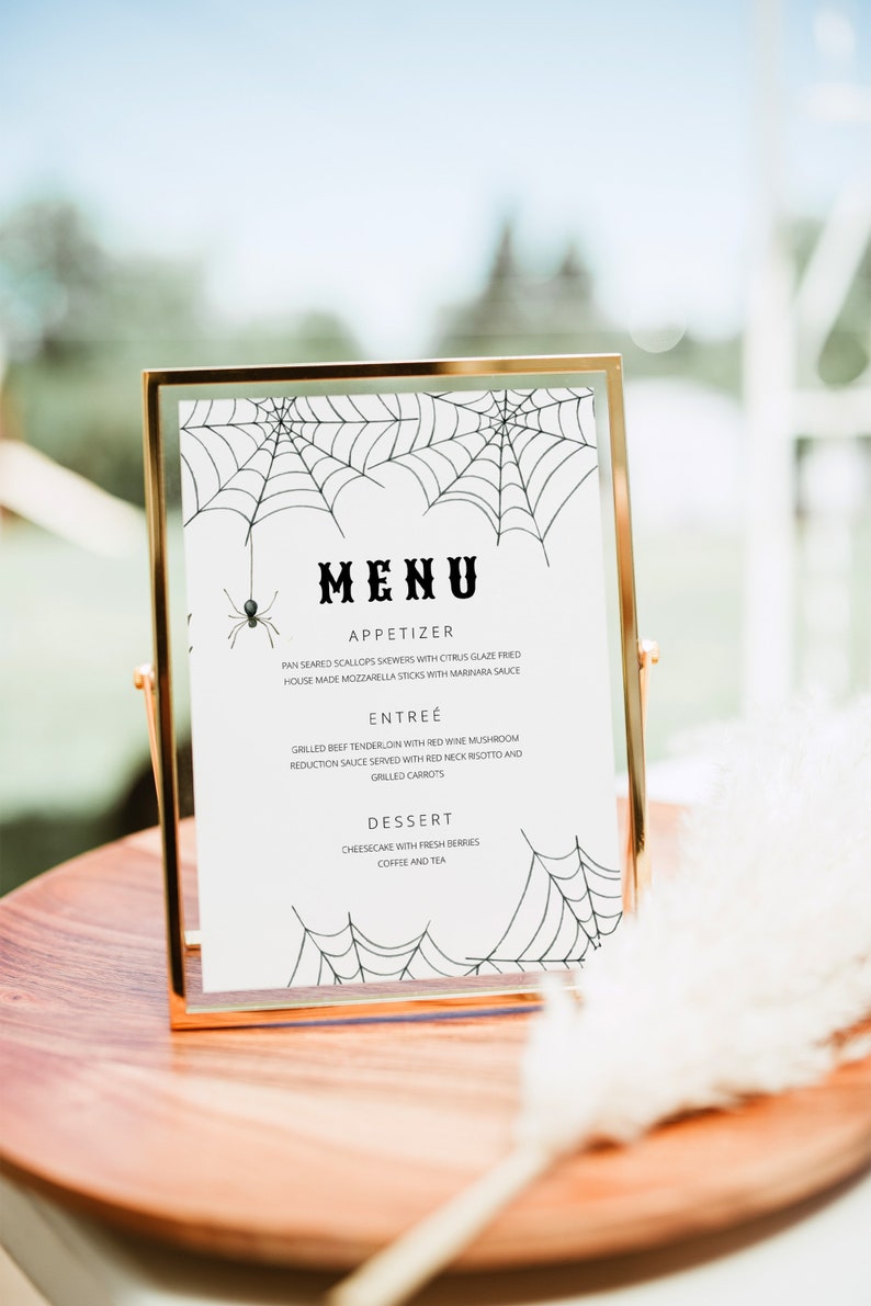 May include: A gold-framed menu with a white background and black spiderweb illustrations. The menu lists "MENU", "APPETIZER", "PAN SEARED SCALLOPS SKEWERS WITH CITRUS GLAZE FRIED", "HOUSE MADE MOZZARELLA STICKS WITH MARINARA SAUCE", "ENTRE&Eacute;", "GRILLED BEEF TENDERLOIN WITH RED WINE MUSHROOM", "REDUCTION SAUCE SERVED WITH RED NECK RISOTTO AND", "GRILLED CARROTS", "DESSERT", "CHEESECAKE WITH FRESH BERRIES", "COFFEE AND TEA".